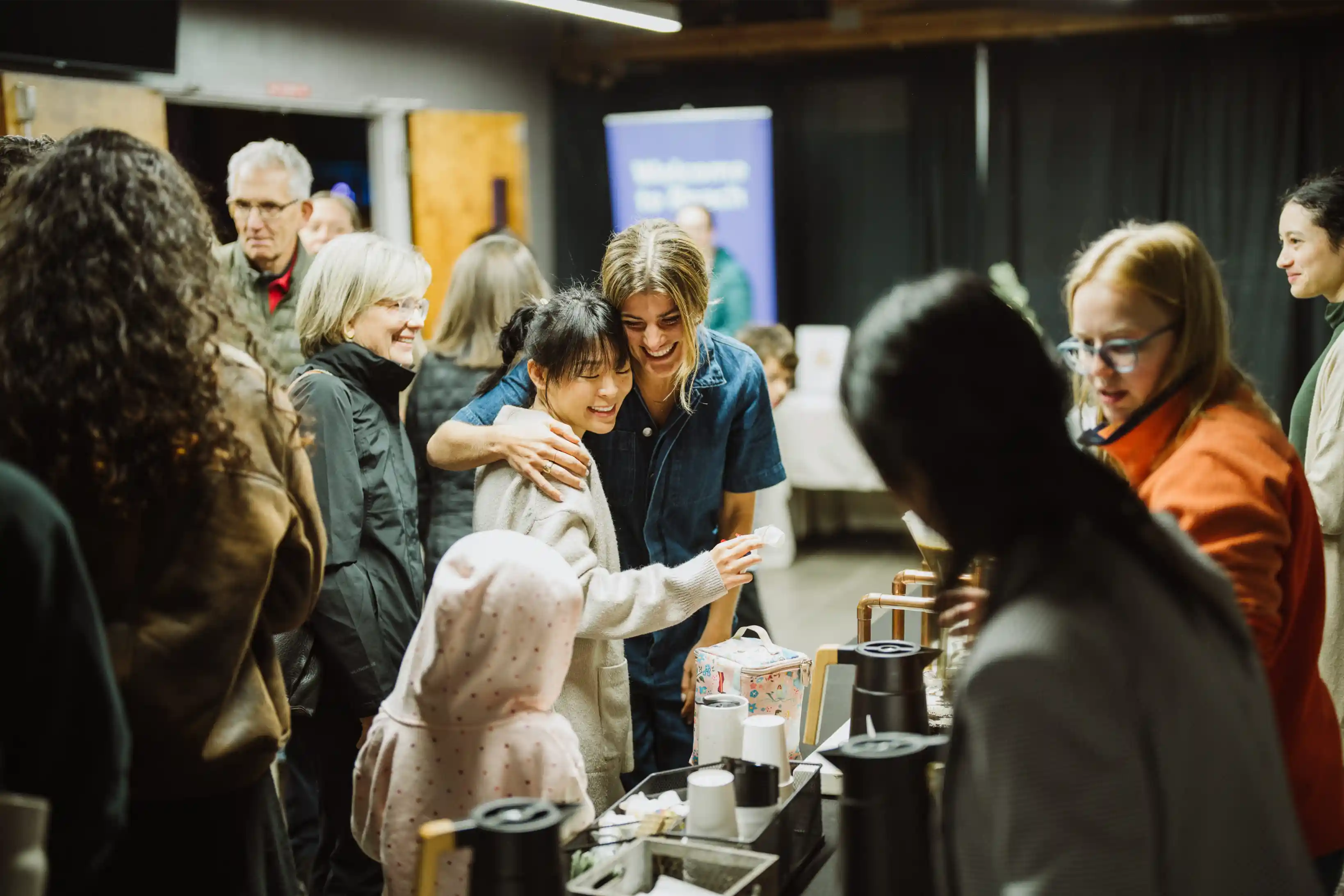 Friends smiling and embracing at a coffee station during an event.