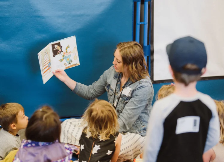 Woman sitting on the floor reading a picture book to a group of attentive children.