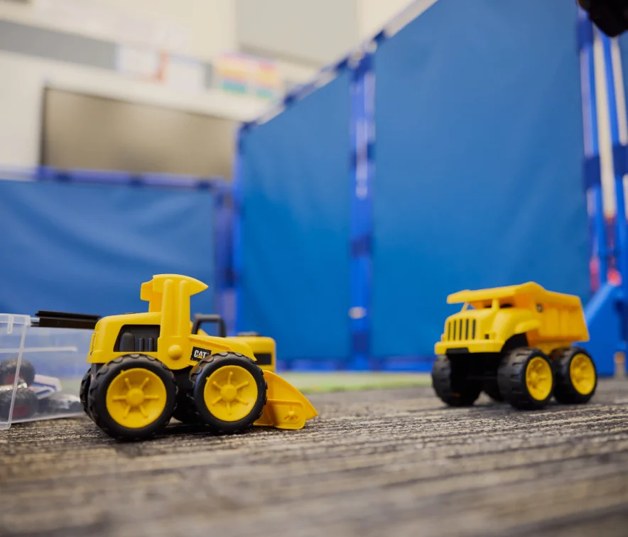 Two yellow toy construction trucks on a carpeted floor with a blue playpen in the background.