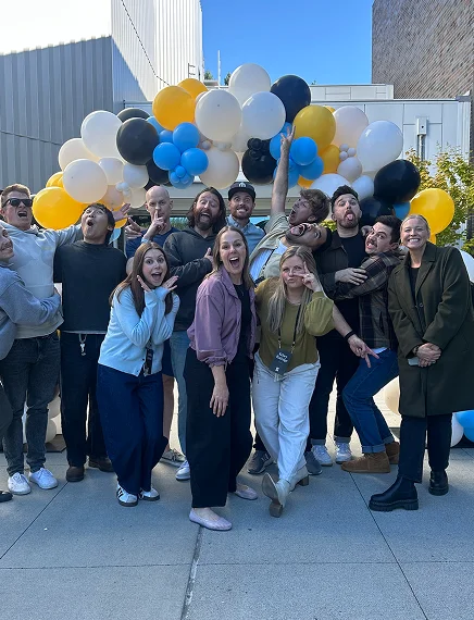 Group of friends posing joyfully outdoors in front of a colorful balloon arch under a clear blue sky.