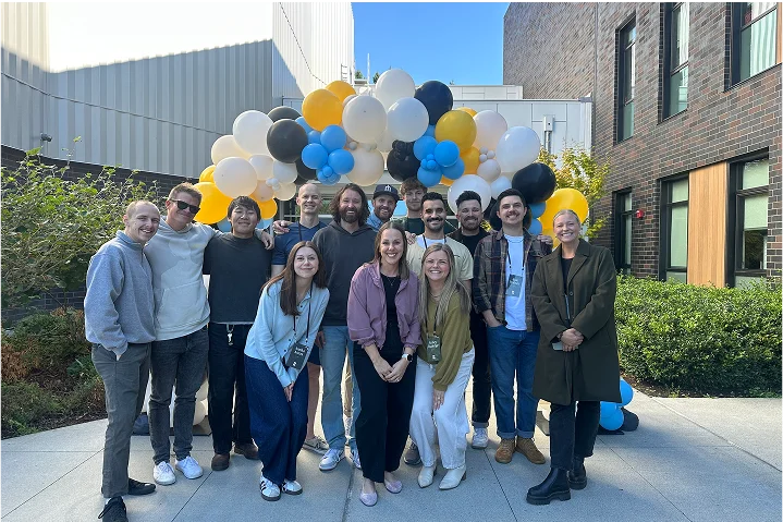Group of twelve people standing outdoors in front of a yellow, white, black, and blue balloon arch, smiling at the camera.