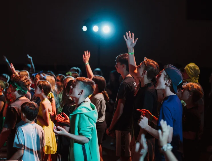 Group of young people standing indoors with hands raised, illuminated by bright stage lights in the background.