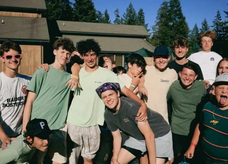 Group of teenage boys smiling and posing together outdoors near wooden buildings and tall pine trees on a sunny day.