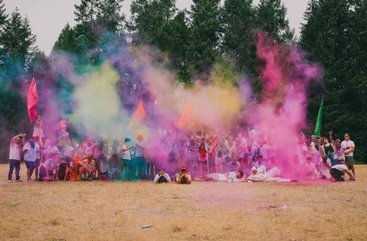 Large group of people outdoors playing with colorful powder creating clouds of pink, yellow, and blue in a grassy area with trees in the background.