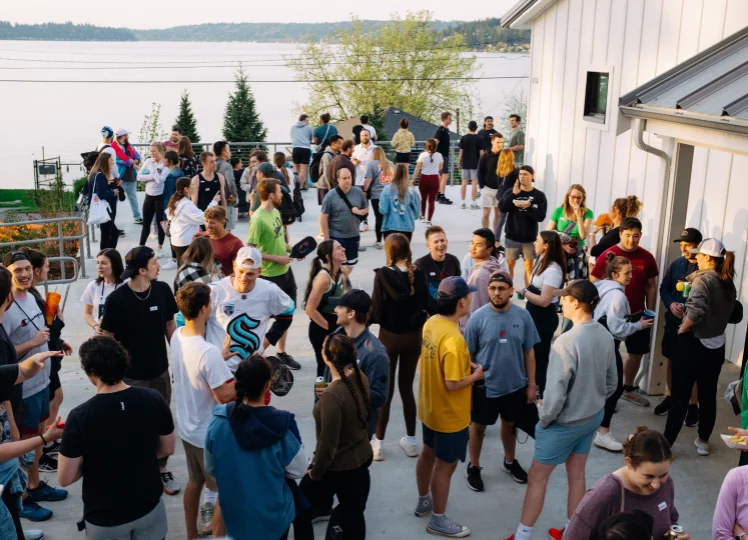 Large group of young people socializing outdoors near a body of water with trees in the background.