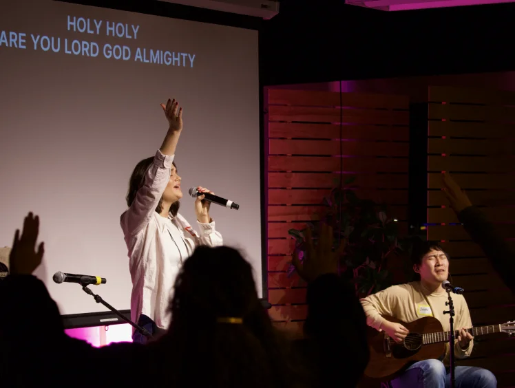 Woman singing with microphone and raised hand on stage while man plays guitar and sings during worship service.