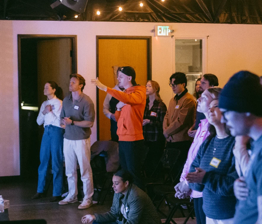 Group of people standing and one person sitting indoors, some with eyes closed and hands clasped, appearing engaged in a solemn or reflective activity.