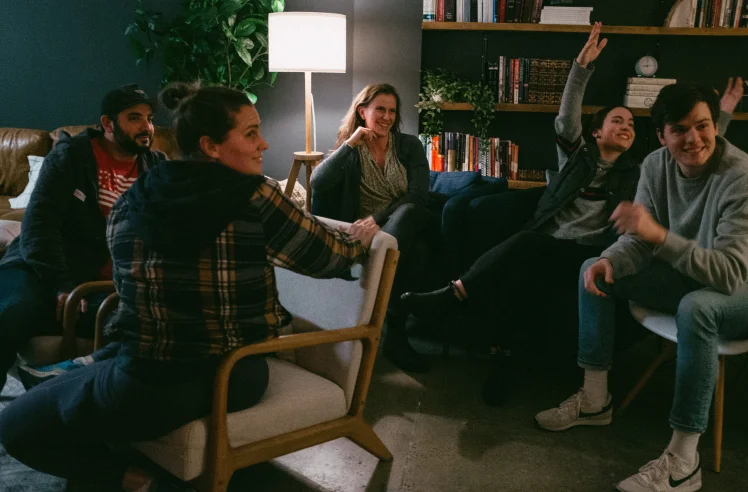 Five people sitting and smiling in a cozy room with bookshelves, a lamp, and plants.