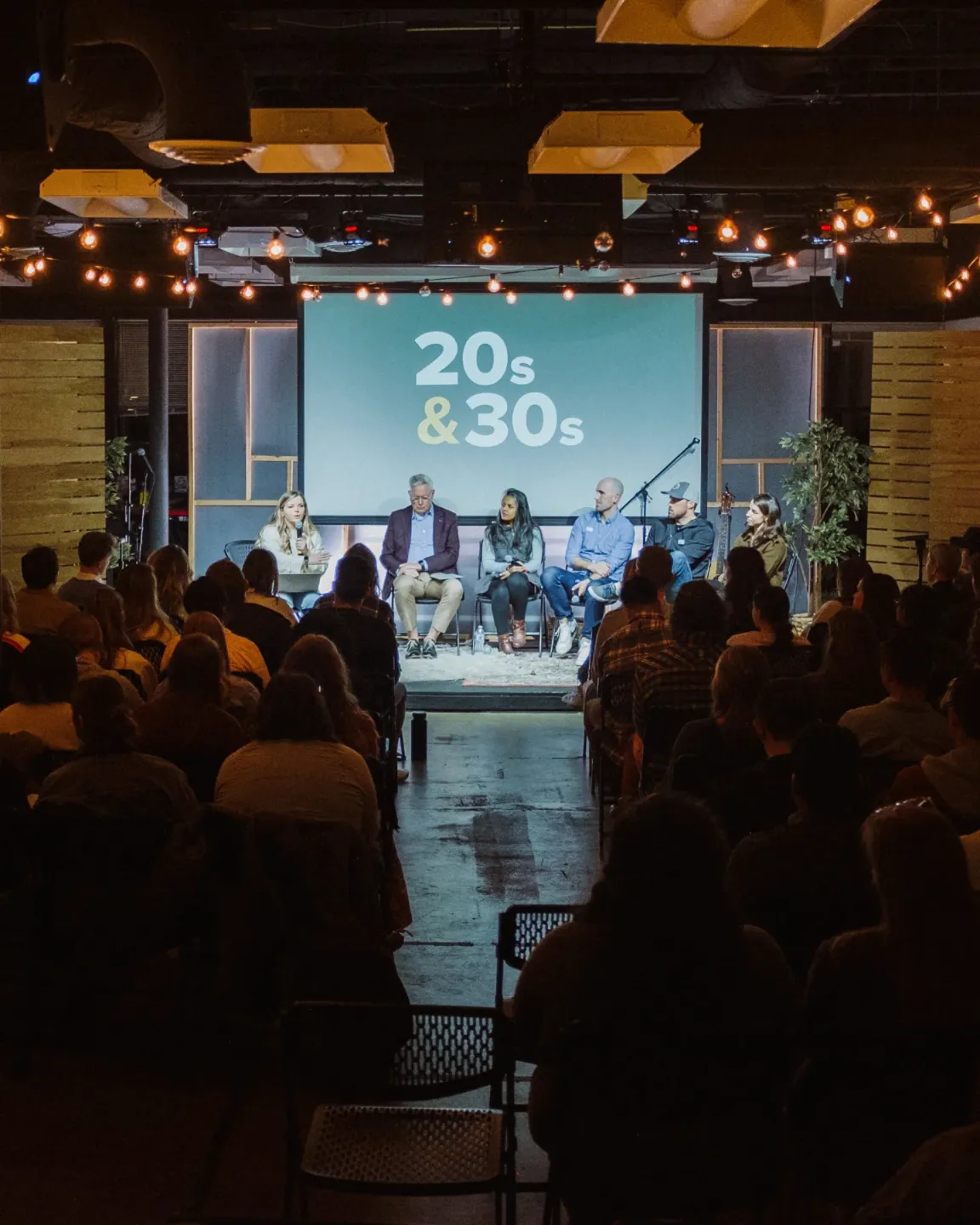 Panel of six speakers seated on stage in front of a screen displaying '20s & 30s' with an audience watching.