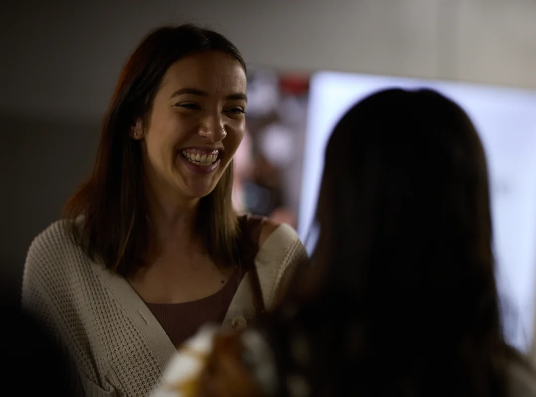 Smiling woman with brown hair talking to another person indoors.