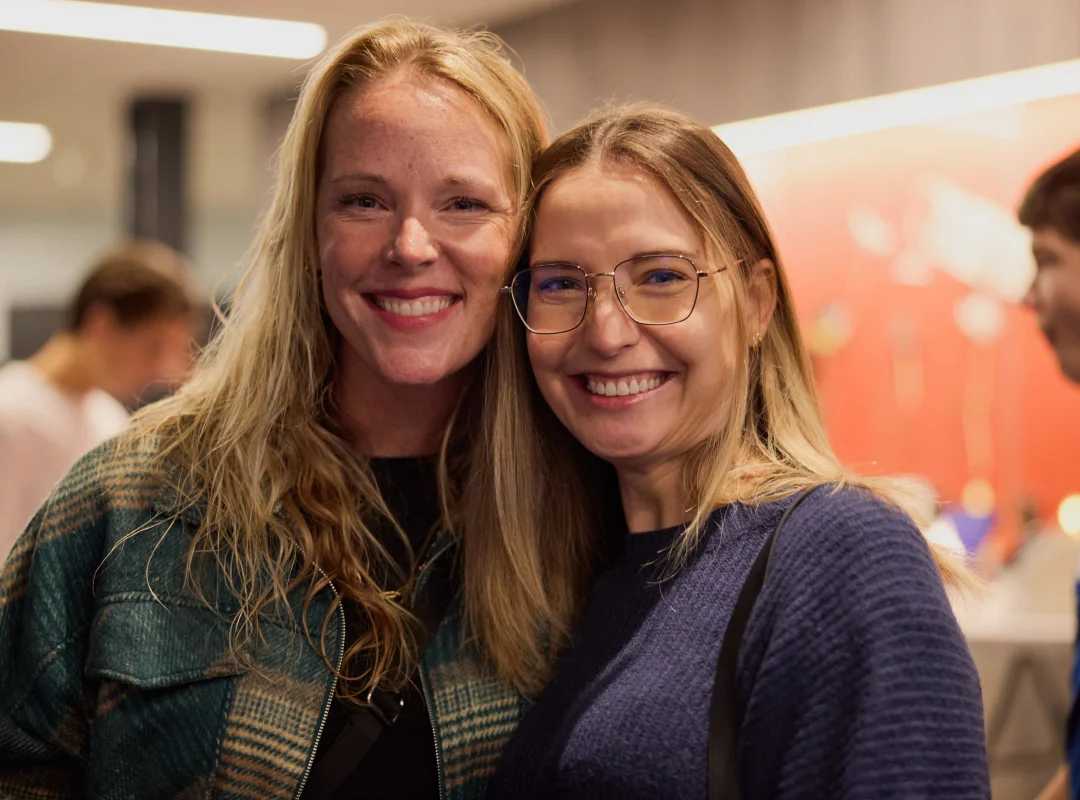 Two smiling women with blonde hair posing closely indoors, one wearing glasses and a blue sweater, the other in a green plaid jacket.