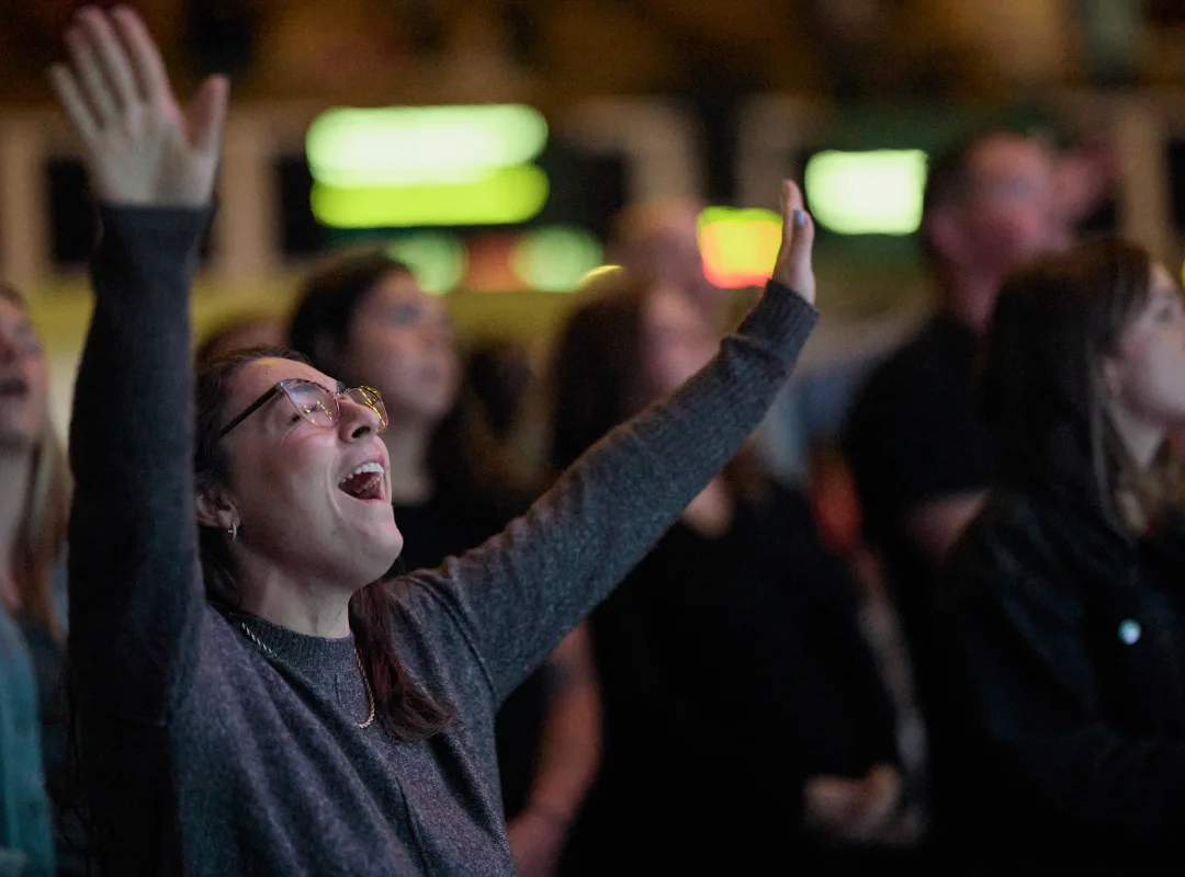 Woman wearing glasses with eyes closed, raising hands in worship amid a crowd.
