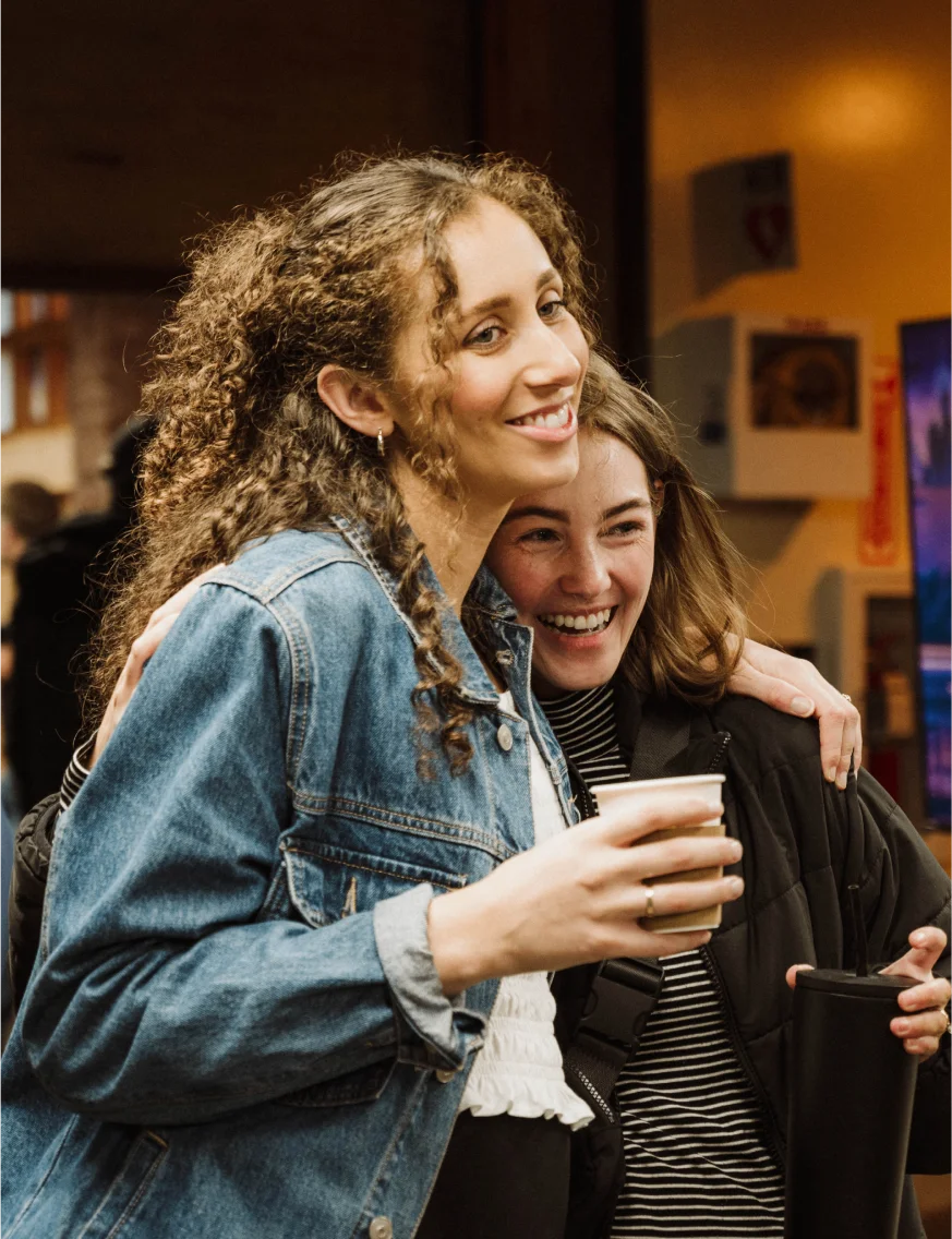 Two young women smiling and hugging, each holding a drink cup indoors.