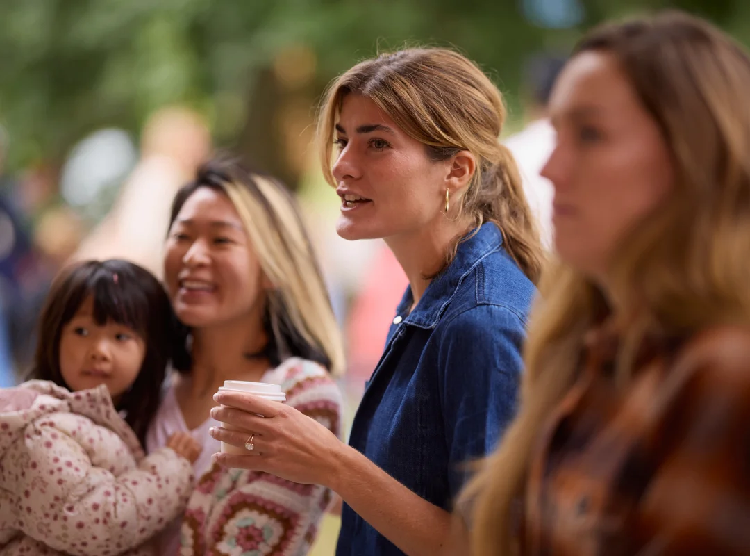 Three women and a young girl outdoors, one woman holding a cup and speaking.