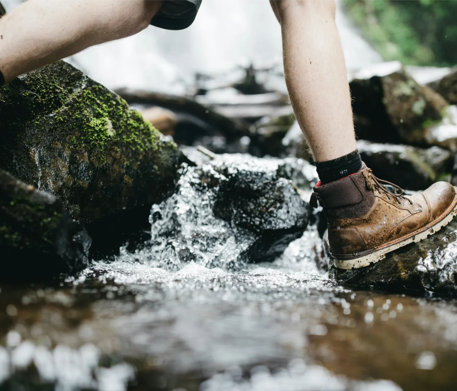 Close-up of a hiker's legs and muddy boots stepping on rocks while crossing a flowing stream.