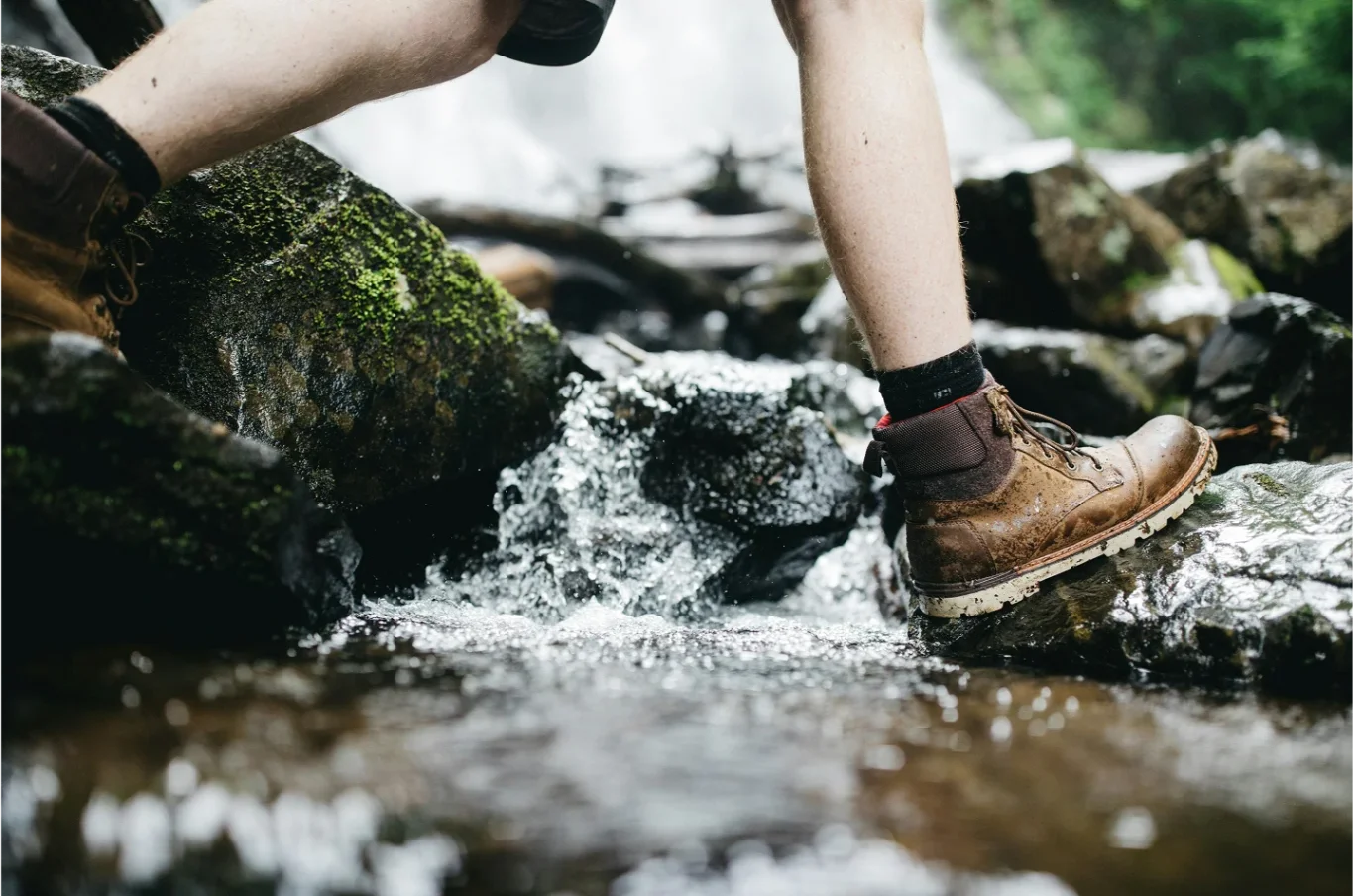 Person wearing brown hiking boots stepping on wet rocks beside a flowing stream.