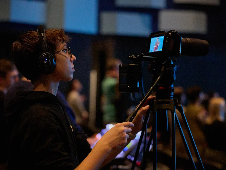 Young videographer wearing headphones and glasses recording an event with a camera on a tripod in a dimly lit indoor setting.