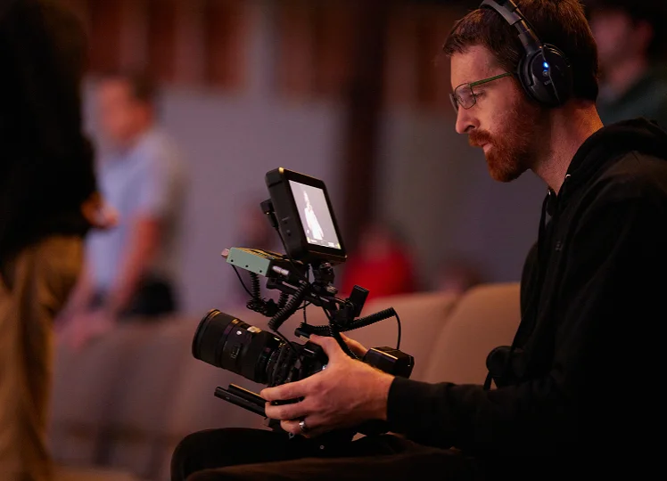 Man wearing glasses and headphones operating a professional video camera with a monitor indoors.