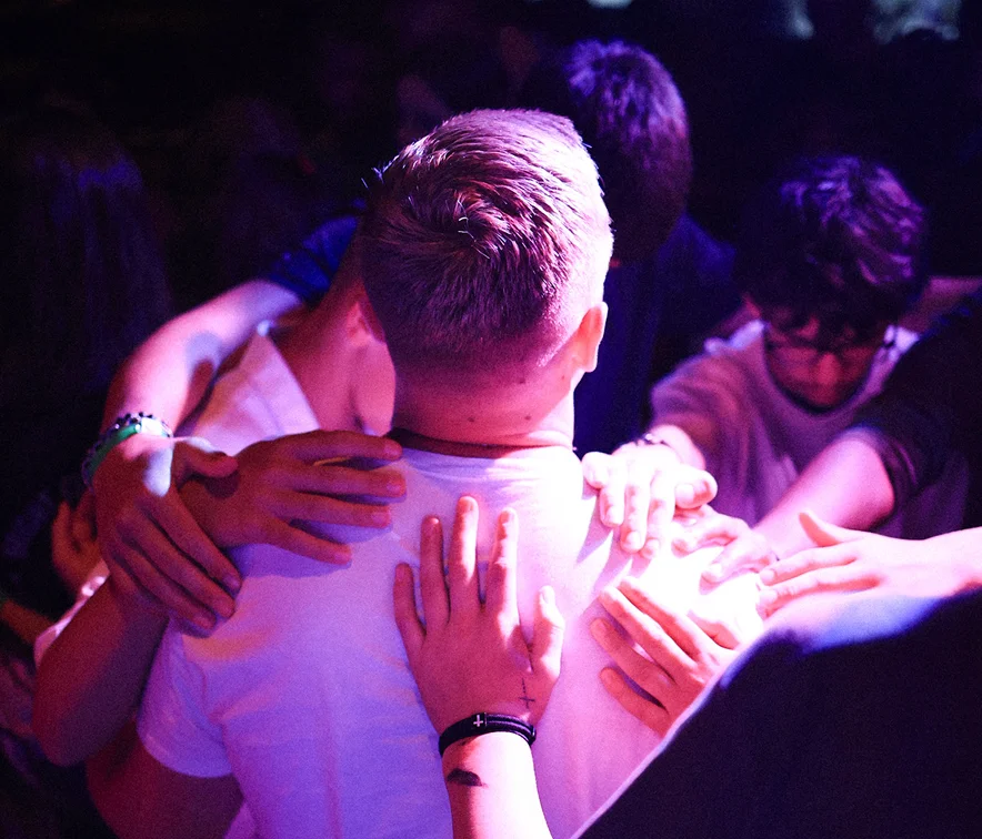 Group of people standing closely with hands placed on a person's back under purple lighting.