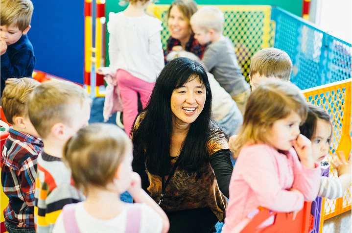 Teacher smiling and interacting with a group of young children in a colorful play area.