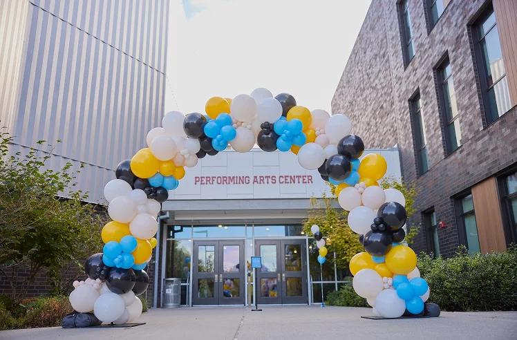 Colorful balloon arch of white, yellow, black, and blue balloons outside the entrance of a building labeled Performing Arts Center.