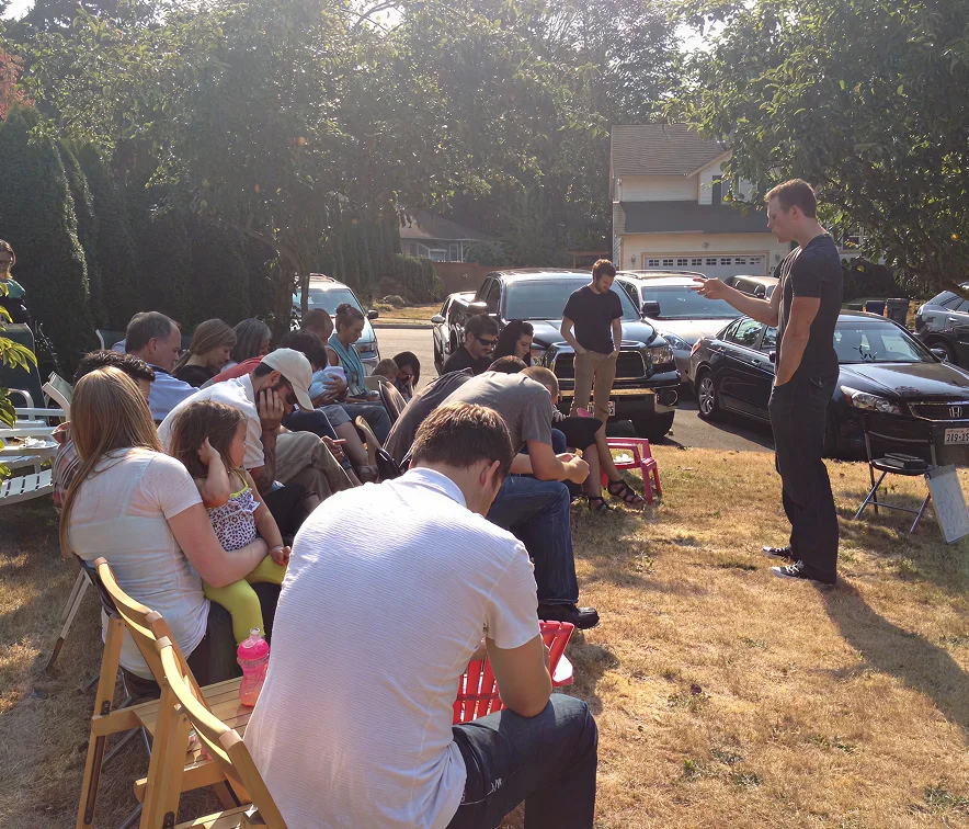 Group of people seated outdoors on folding chairs, listening to a standing man speaking, with cars and a house in the background.