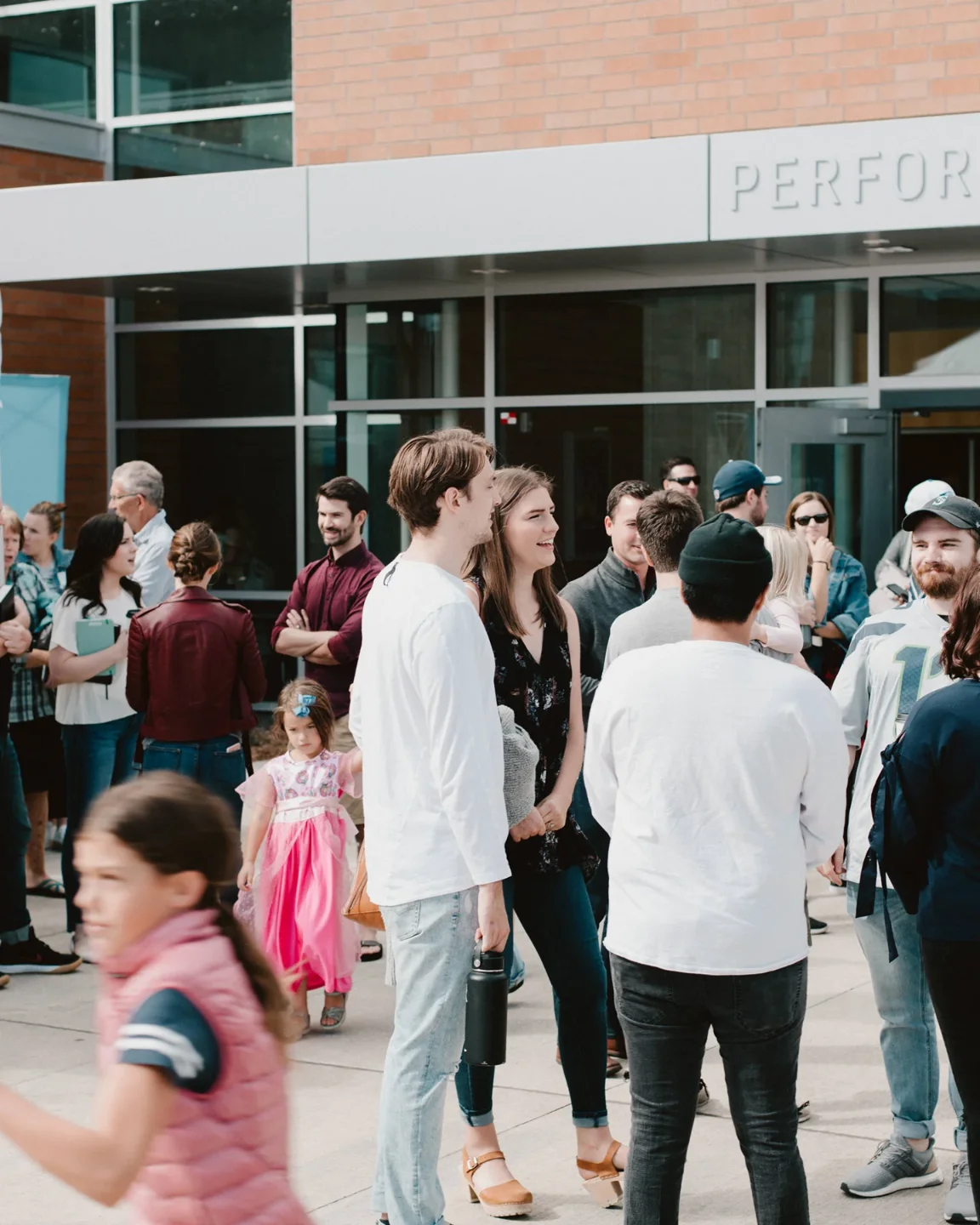 Group of people socializing outside a building with glass doors and brick walls, including adults and children.