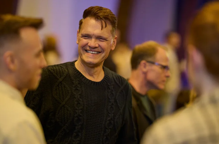 Smiling man in a black sweater chatting with two other men at a social event.