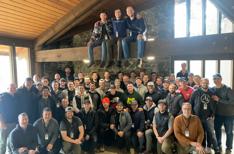 Large group of men posing inside a rustic room with wooden ceiling and stone wall, some sitting on a beam above.