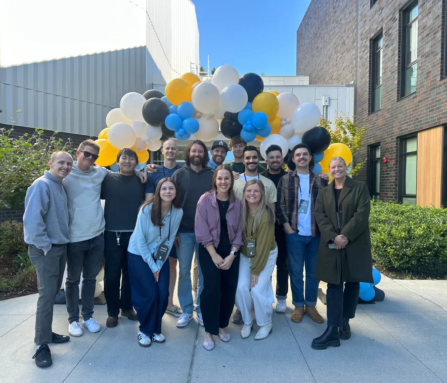 Group of smiling people posing in front of a balloon arch with white, yellow, black, and blue balloons outside on a sunny day.