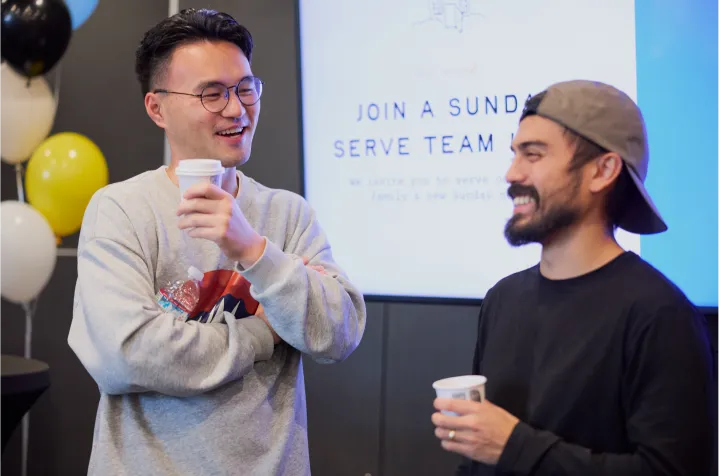 Two men smiling and holding coffee cups while conversing indoors with balloons and a sign in the background.