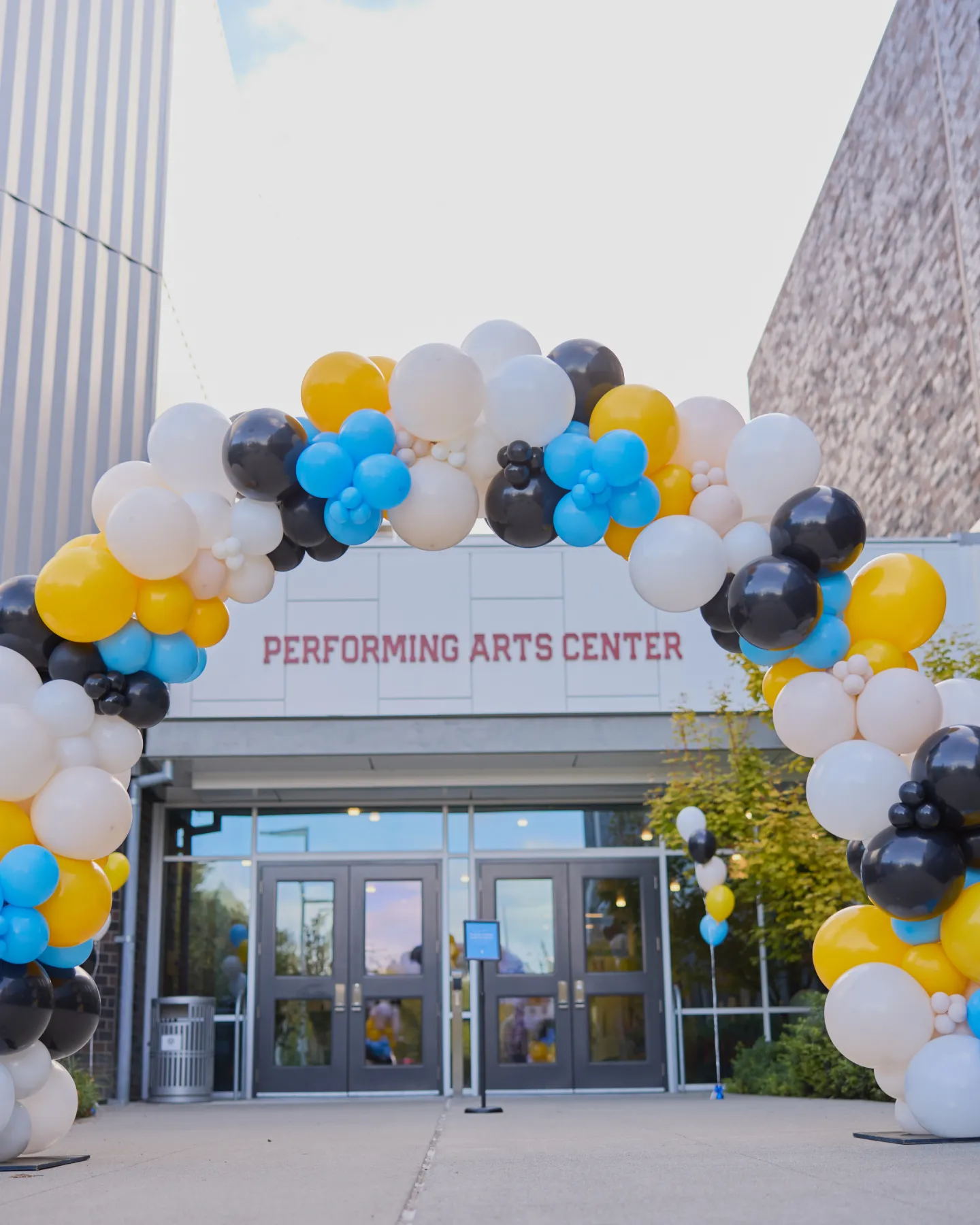 Colorful balloon arch in yellow, black, blue, and white at the entrance of a building labeled Performing Arts Center.