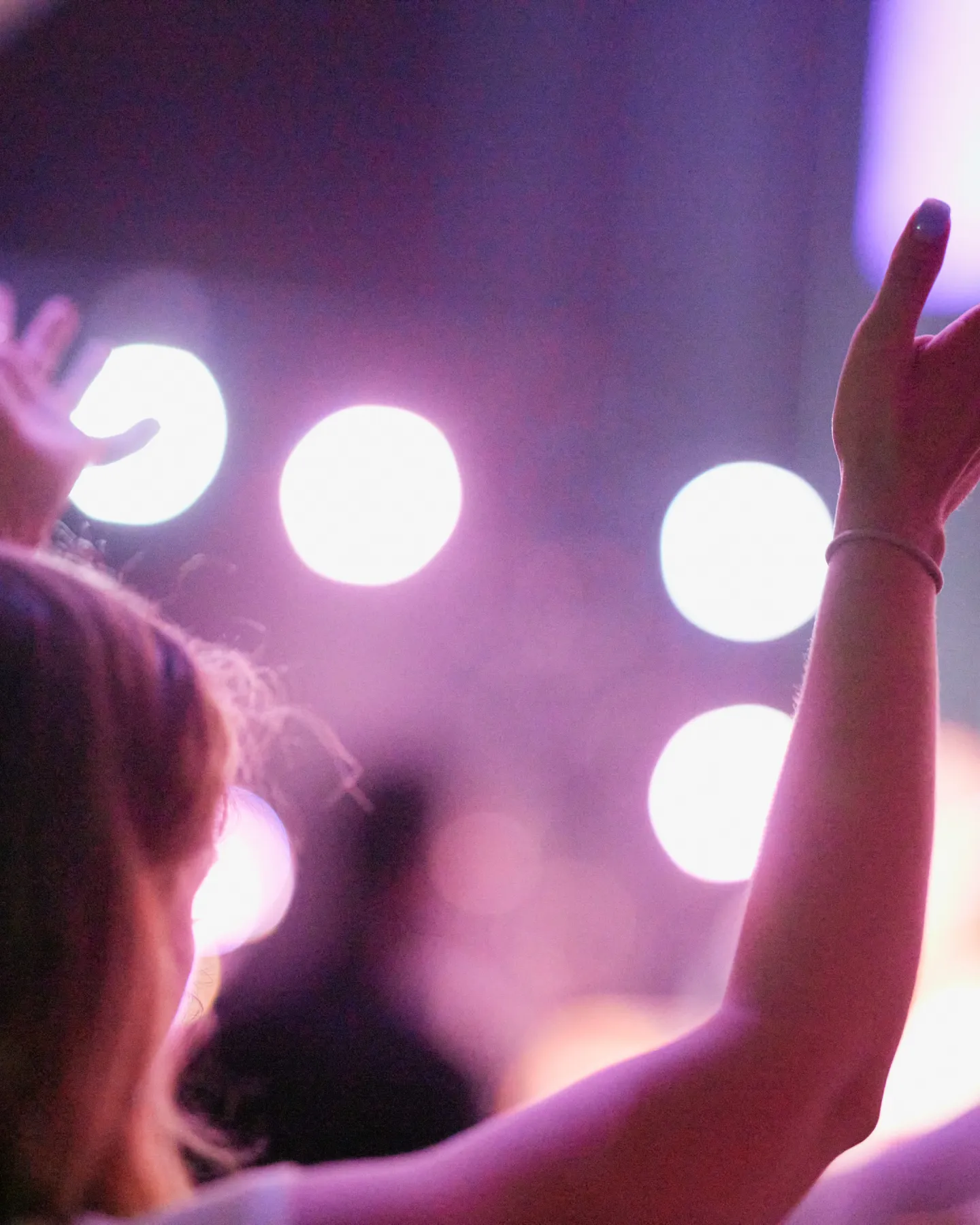 Person raising both hands with blurred purple and white stage lights in the background during a worship event.