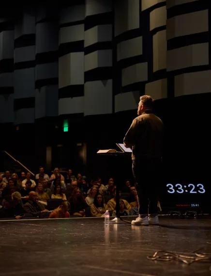 Man speaking on a stage with a laptop and open book, addressing a seated audience in a dark auditorium.