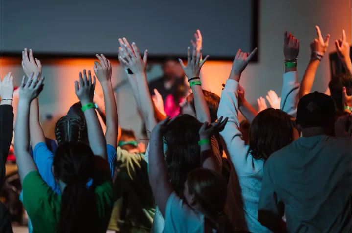 Group of children raising their hands enthusiastically at an indoor event with colorful lighting.