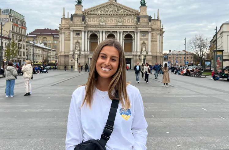 Young woman smiling in front of a historic ornate building in a busy urban square.
