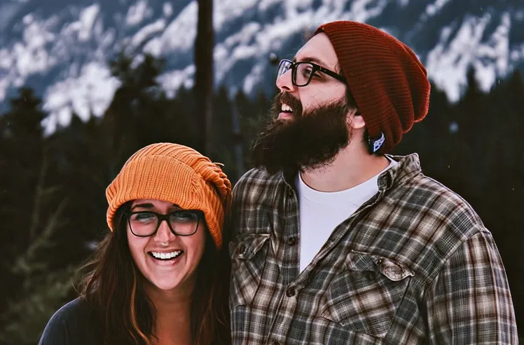 Smiling couple wearing glasses and knit beanies standing outdoors with snowy mountains in the background.