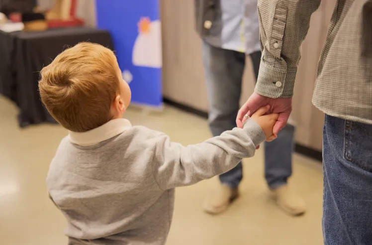 Young child reaching up and holding an adult's hand indoors.