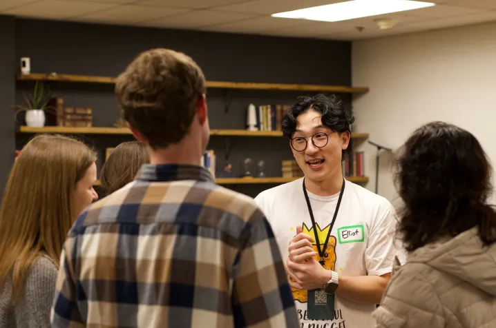 A young man named Elliot, wearing glasses and a white t-shirt, speaks animatedly to a small group of people in a cozy room with bookshelves.