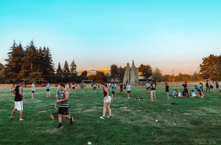Group of people playing games and socializing on a grassy field at sunset with trees and sculptures in the background.