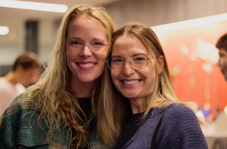 Two women smiling closely together at an indoor gathering with blurred people in the background.
