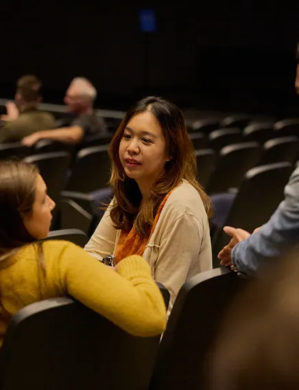 Woman with long hair in a beige sweater listening attentively to another woman in a yellow sweater in a dimly lit auditorium.