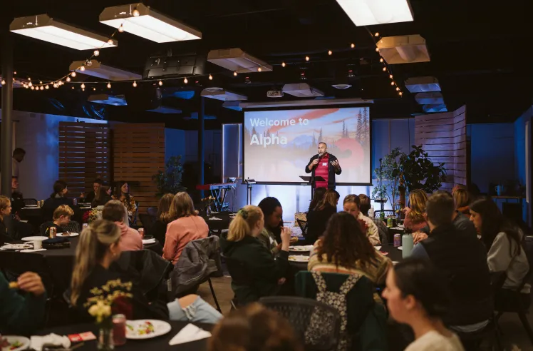 Man speaking on stage in front of an audience seated at tables with a screen reading 'Welcome to Alpha' behind him.