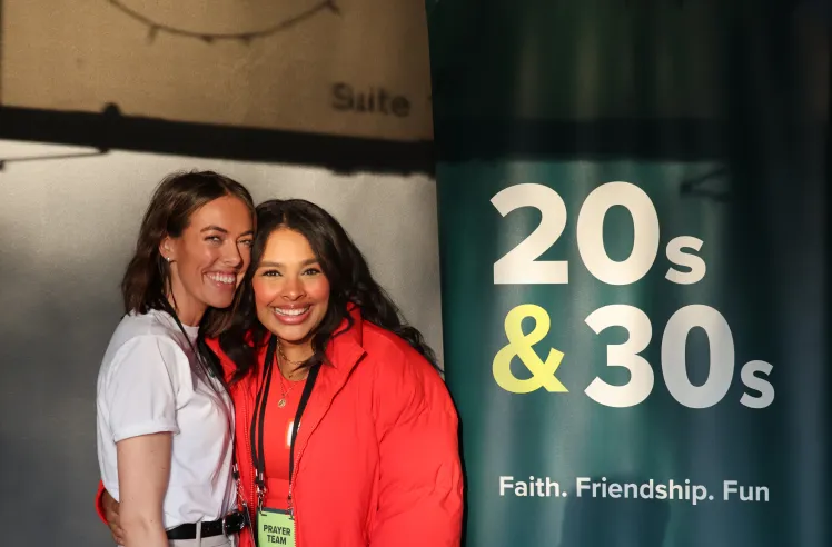 Two smiling women standing close together at a 20s and 30s event with a banner reading '20s & 30s Faith. Friendship. Fun.'
