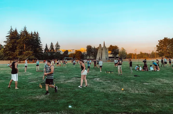 People playing various outdoor games and socializing on a large green field with trees and a sculpture in the background during sunset.