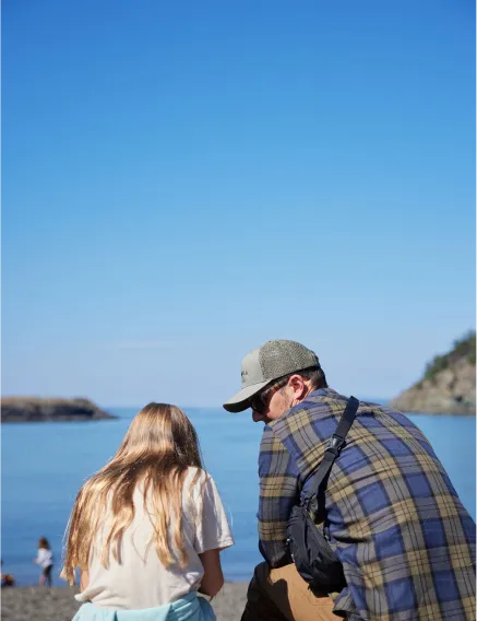 Couple sitting on a beach facing the ocean on a sunny day, with a clear blue sky overhead.