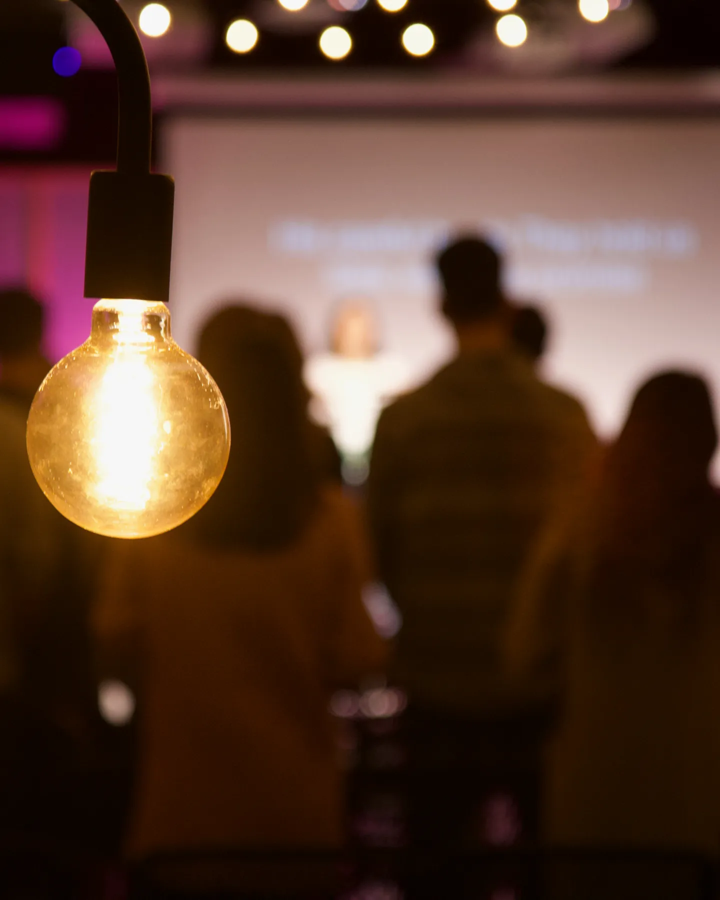 Glowing hanging light bulb in focus with a blurred crowd of people and string lights in the background.