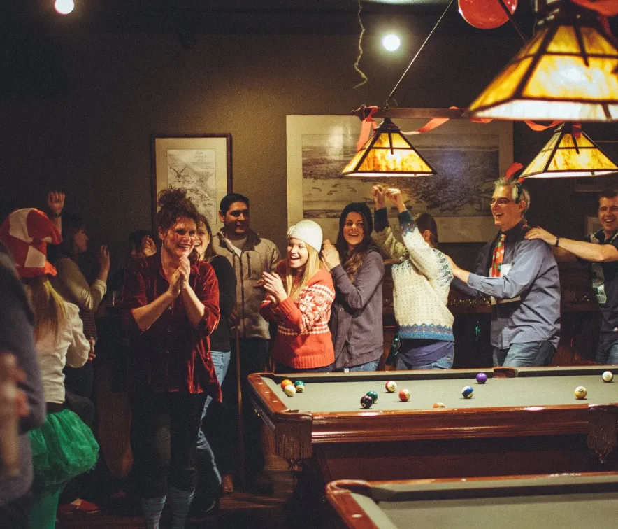 Group of people joyfully celebrating indoors near a pool table with festive decorations and warm lighting.