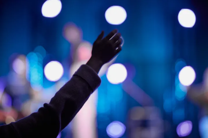 Silhouette of a raised hand in a worship setting with blue stage lights in the background.