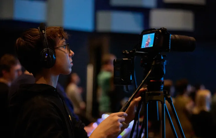 Young camera operator wearing headphones filming an event with a professional video camera on a tripod.
